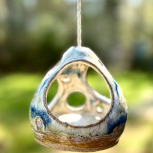 Könnte beinhalten: Handgefertigter Vogelhaus-Futternapf aus Keramik mit einzigartigem, offenem Design. Der Futterspender wird an einer Naturfaserschnur aufgehängt. Er hat eine cremefarbene Basis mit blauen und braunen Akzenten und mehrere kreisförmige Öffnungen.