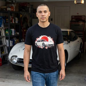 May include: A black short-sleeved t-shirt featuring a graphic print of a white vintage sports car centred in front of a large red circle with white paint splatter effects. The design is positioned on the chest area of the garment.