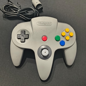 May include: A gray Nintendo 64 controller with a black cable. The controller features a directional pad, a red start button, green and blue buttons, and yellow buttons. The Nintendo logo is visible on the top.