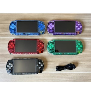 May include: A collection of five Sony PSP handheld gaming consoles in various colors: blue, purple, red, green, and black. Each console features a screen, buttons, and a D-pad. A black charging cable is also included.