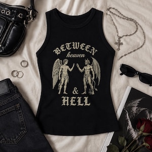 May include: A black sleeveless tank top featuring a beige graphic print of an angel and a devil facing each other. The text reads BETWEEN heaven & HELL in a gothic font. The design is centred on the chest of the garment, which has a crew neckline.