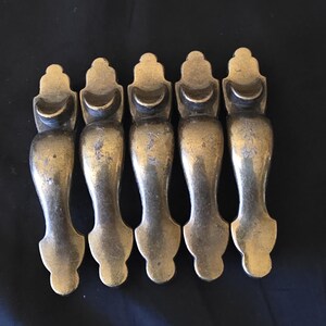 May include: Five matching antique brass drawer pulls arranged vertically. Each handle features a curved, elongated body with a decorative, rounded top finial and a smooth, tapered base. The metal surface displays a weathered, aged patina finish.