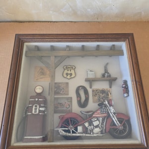 Pode incluir: Uma caixa de sombra emoldurada com um tema de motocicleta vintage. A exibição inclui uma motocicleta vermelha com um design de bandeira americana, uma bomba de gasolina e sinalização da Rota 66. A moldura é marrom.