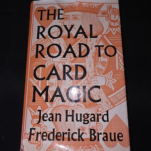 May include: A vintage book titled "The Royal Road to Card Magic" by Jean Hugard and Frederick Braue. The cover is orange and white, featuring a playing card king illustration and text in a bold, serif font. The book is set against a black background.