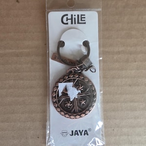 May include: A bronze-colored keychain with a circular pendant featuring a decorative design. The keychain is packaged in a clear plastic sleeve with the word "CHILE" and the brand name "JAYA" printed on the card.