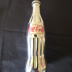 May include: A silver-colored Coca-Cola bottle with the brand name in red script. The bottle has a classic shape with a silver cap and the words "Enjoy Coca-Cola Trademark" printed on the front.