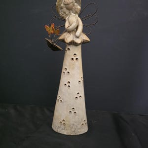 May include: A tall, beige ceramic figurine of a figure wearing a wide-brimmed hat and a flared skirt. The figure has wire wings, holds a small black umbrella with two orange butterflies attached, and features a pattern of small, triple-dot cutouts.