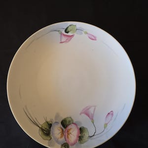 May include: A round white ceramic bowl featuring hand-painted floral motifs in shades of pink, blue, and green. The design includes delicate blossoms and leaves arranged along the inner rim, with a smooth, glossy finish and a clean, simple edge.
