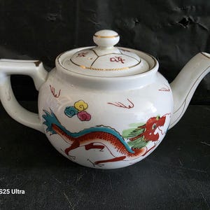 May include: A white porcelain teapot featuring a hand-painted red and blue dragon motif along the body. The lid includes a small knob and gold-toned trim, with red calligraphic characters. The spout and handle are white with gold-toned accents.