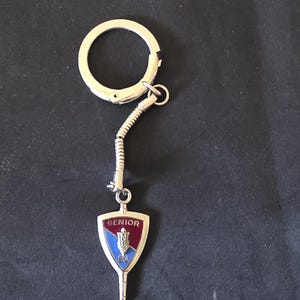 May include: A silver-toned metal keychain featuring a flexible coiled link attached to a shield-shaped pendant. The pendant has a maroon top section with the word SENIOR in white, and a blue bottom section with the letters NB and the number 81.