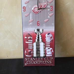 May include: A rectangular wall clock featuring the Detroit Red Wings logo, the text Olympia, and the Stanley Cup trophy. The design includes images of hockey jerseys and championship years, with black clock hands and red and white accents.