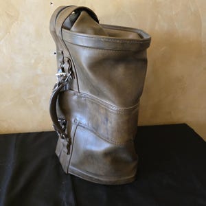 May include: A tall, cylindrical olive green bag with a smooth, matte finish. The bag features a sturdy side handle attached with silver-toned metal hardware and a zippered top closure. The structure is upright with reinforced horizontal stitching.