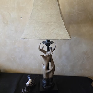 May include: A table lamp featuring a beige, textured, rectangular lampshade and a base sculpted to resemble intertwined brown antlers. The lamp includes a black square base, a central metal rod, and a pull chain with a small antler-shaped toggle.
