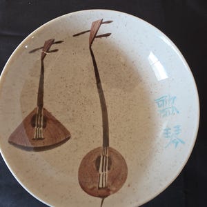 Puede incluir: Un plato de cerámica beige con una superficie moteada. Dos ilustraciones marrones de instrumentos de cuerda están pintadas en el plato. Caracteres japoneses azules en el lado derecho.