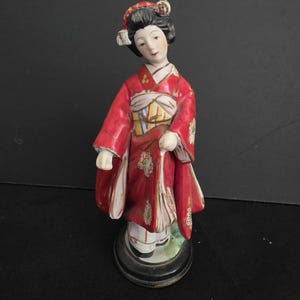May include: A porcelain figurine of a person with black hair styled in an updo, wearing a vibrant red kimono with gold accents and a patterned obi sash. The figure stands on a circular black base, featuring painted floral details on the fabric.