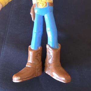 May include: A toy figure of a cowboy, featuring a yellow vest, blue pants, and brown cowboy boots. The boots have a cactus design. The figure is a popular character from a children's animated movie.