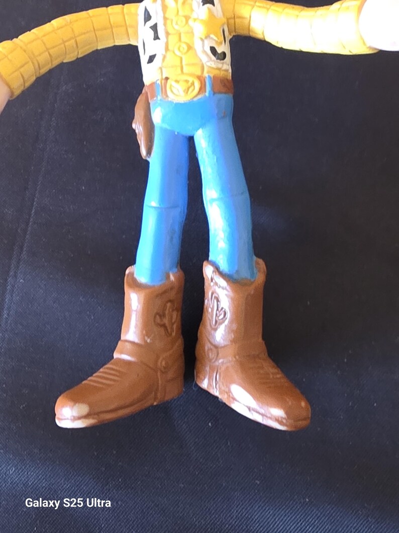 May include: A toy figure of a cowboy, featuring a yellow vest, blue pants, and brown cowboy boots. The boots have a cactus design. The figure is a popular character from a children's animated movie.