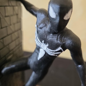K&ouml;nnte beinhalten: Schwarze Actionfigur eines Superhelden mit wei&szlig;en Spinnennetzdetails auf Brust und Maske. Die Figur ist in einer dynamischen Pose vor einer strukturierten Backsteinmauer dargestellt.
