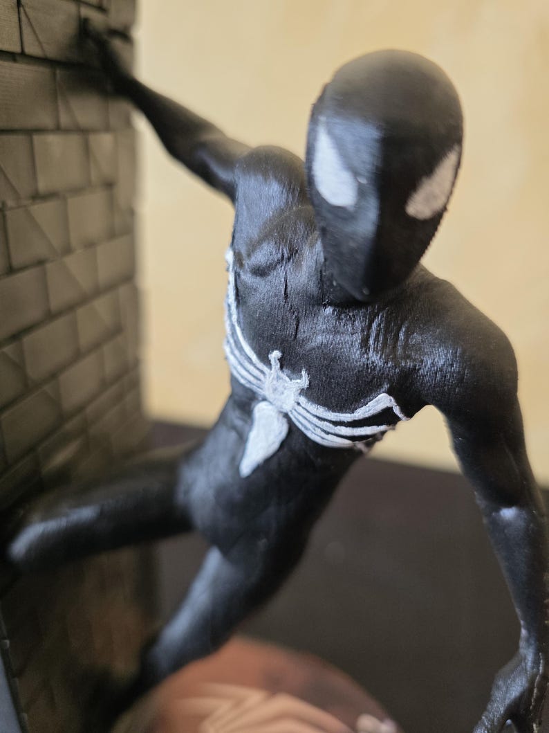 K&ouml;nnte beinhalten: Schwarze Actionfigur eines Superhelden mit wei&szlig;en Spinnennetzdetails auf Brust und Maske. Die Figur ist in einer dynamischen Pose vor einer strukturierten Backsteinmauer dargestellt.
