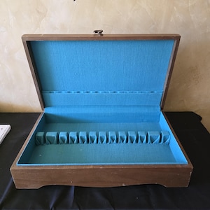 May include: A rectangular wooden storage chest with a hinged lid, featuring a bright turquoise fabric lining. The interior includes a series of molded slots along the base and the lid, designed for organizing flatware or similar items.