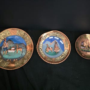 May include: Three circular copper plates featuring embossed borders and central painted scenes. The two larger plates depict figures with llamas in mountainous landscapes, while the smallest plate shows a group of figures, all labeled PERU.