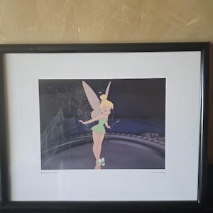 May include: Framed print of Tinkerbell from Peter Pan. The artwork features Tinkerbell in a green dress with blonde hair and fairy wings. The print is in a black frame with a white mat.