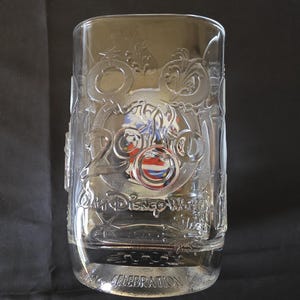 May include: Clear glass tumbler with embossed Disney World 2000 Celebration design. The design includes Mickey Mouse ears, a carriage, and the words "Walt Disney World" and "Celebration". The glass is filled with a clear liquid.