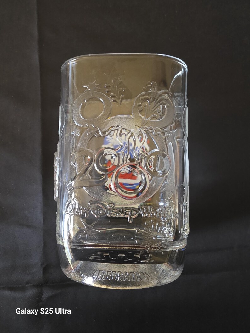 May include: Clear glass tumbler with embossed Disney World 2000 Celebration design. The design includes Mickey Mouse ears, a carriage, and the words "Walt Disney World" and "Celebration". The glass is filled with a clear liquid.