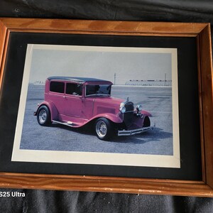 May include: A framed photograph of a vintage pink hot rod car with a black roof and wire wheels, parked on an asphalt surface. The image is enclosed in a rectangular wooden frame with a black mat border, showcasing the classic automobile design.