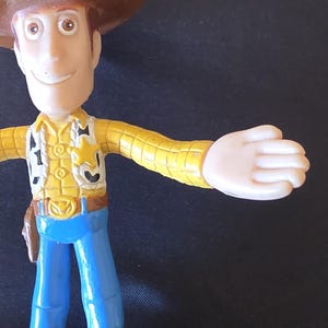 May include: A plastic toy figure of a cowboy character, wearing a brown hat, yellow shirt, blue pants, and a sheriff's badge. The figure has outstretched arms and a friendly expression. The toy is a collectible item.