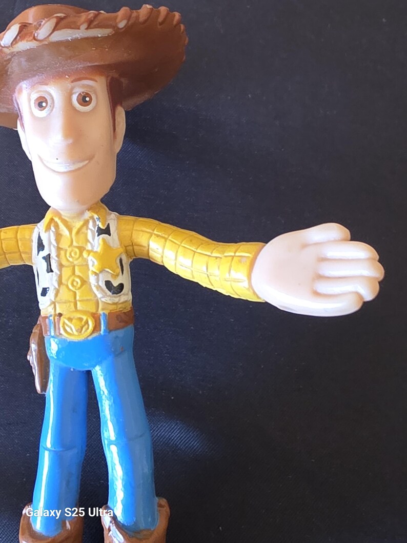 May include: A plastic toy figure of a cowboy character, wearing a brown hat, yellow shirt, blue pants, and a sheriff's badge. The figure has outstretched arms and a friendly expression. The toy is a collectible item.