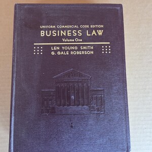 Könnte beinhalten: Ein dunkelviolettes Hardcover-Buch mit dem Titel "BUSINESS LAW Volume One" mit geprägtem Text und einer Gebäudeillustration. Das Buch ist Teil der Uniform Commercial Code Edition.