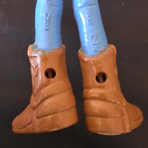 May include: Close-up of the lower legs and boots of a Woody action figure. The figure's legs are light blue, and the boots are brown with a textured design. The boots have a hole in the center. The text "Disney Pixar C-TM" is visible on the leg.