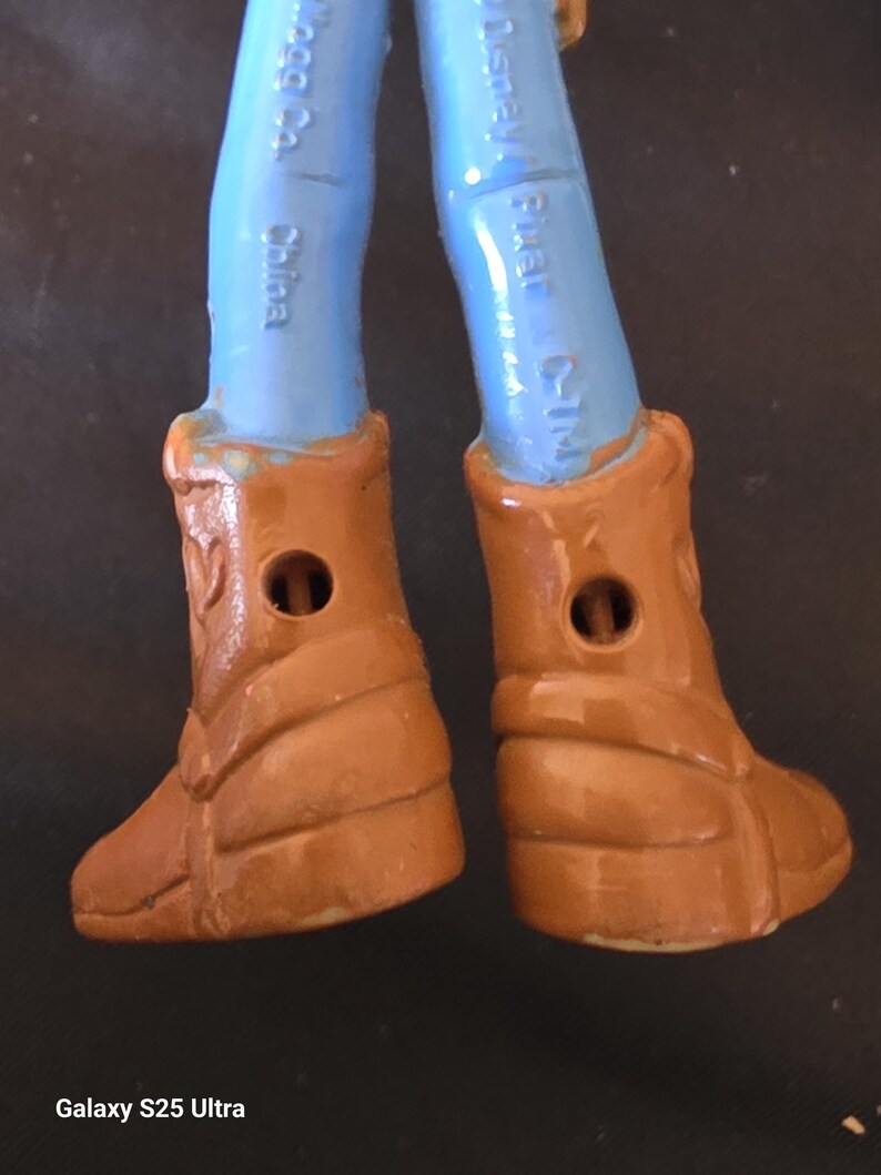 May include: Close-up of the lower legs and boots of a Woody action figure. The figure's legs are light blue, and the boots are brown with a textured design. The boots have a hole in the center. The text "Disney Pixar C-TM" is visible on the leg.