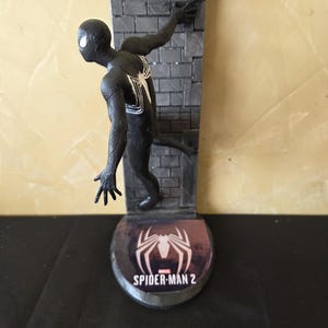 K&ouml;nnte beinhalten: Eine schwarze Spider-Man-Figur mit wei&szlig;em Spinnenemblem auf dem R&uuml;cken, die an einer grauen Backsteinmauer haftet. Der Sockel zeigt das Spider-Man 2-Logo in Wei&szlig; auf weinrotem Hintergrund. Die Figur ist in einer dynamischen Pose.