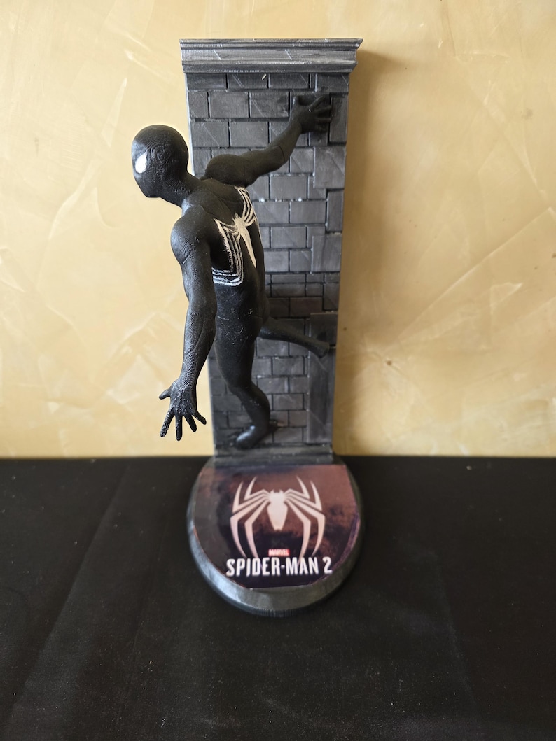 K&ouml;nnte beinhalten: Eine schwarze Spider-Man-Figur mit wei&szlig;em Spinnenemblem auf dem R&uuml;cken, die an einer grauen Backsteinmauer haftet. Der Sockel zeigt das Spider-Man 2-Logo in Wei&szlig; auf weinrotem Hintergrund. Die Figur ist in einer dynamischen Pose.
