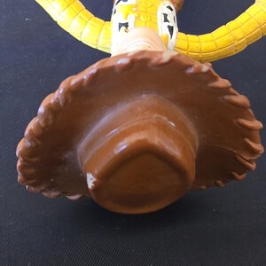 May include: A close-up of a Woody action figure from the Toy Story franchise. The figure is wearing a brown cowboy hat, a yellow shirt, blue pants, and a brown vest. The figure's face is visible, with a painted expression.