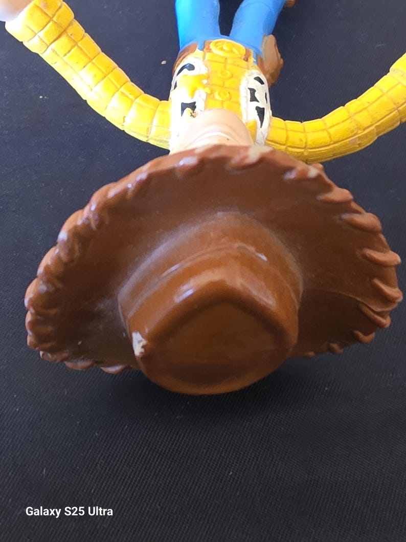 May include: A close-up of a Woody action figure from the Toy Story franchise. The figure is wearing a brown cowboy hat, a yellow shirt, blue pants, and a brown vest. The figure's face is visible, with a painted expression.