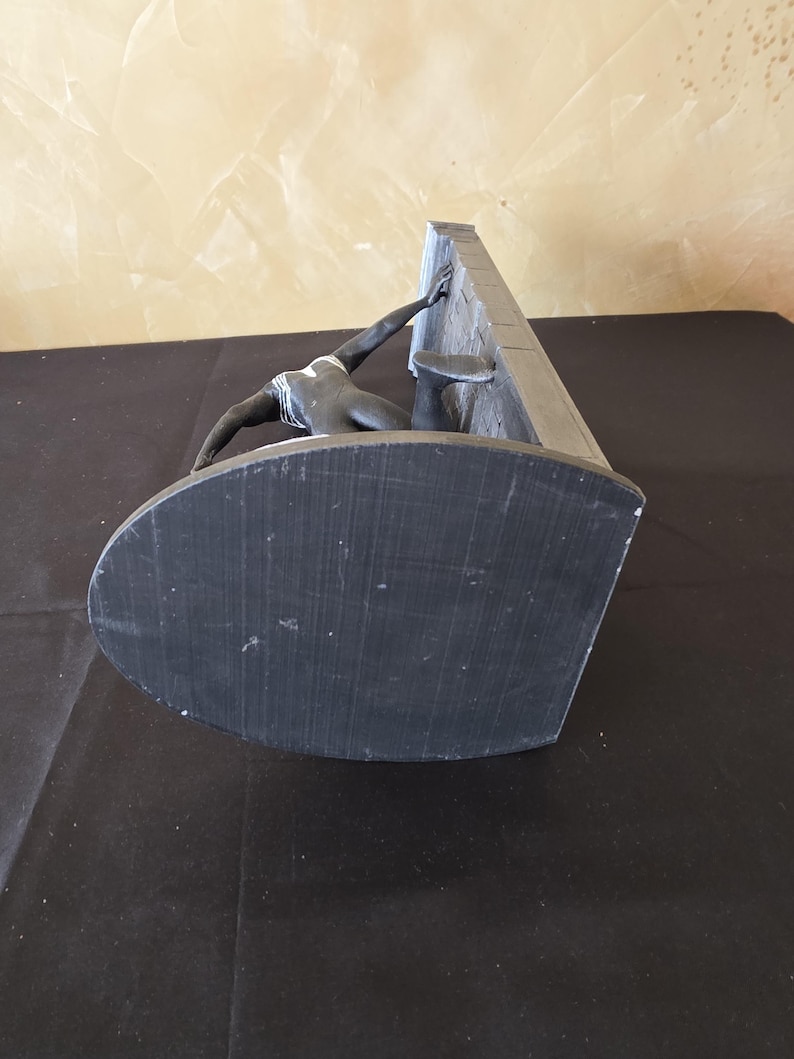 K&ouml;nnte beinhalten: Eine dunkelgraue Skulptur, die eine Figur darstellt, die nach einer Wand greift. Die Figur kniet, ein Arm ausgestreckt. Die Skulptur hat einen abgerundeten Sockel und steht vor einem neutralen Hintergrund.