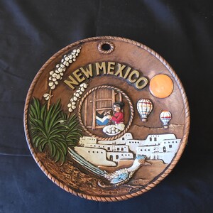 May include: A brown ceramic plate with a raised relief design featuring the text "NEW MEXICO". The plate depicts a Southwestern scene with a roadrunner, hot air balloons, and a Native American figure.