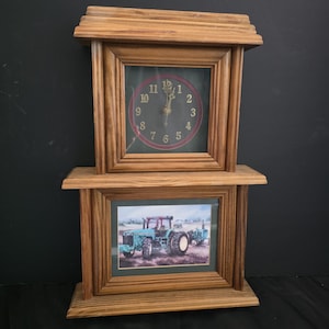 May include: A wooden wall clock featuring a square clock face with gold numbers one through twelve on a dark background, positioned above a rectangular framed print of a blue tractor in a field, both encased in a light brown wooden frame structure.