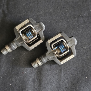 May include: A pair of clipless bicycle pedals featuring a black composite body, silver metal cage components, and a prominent blue coiled tension spring mechanism. The pedals include threaded steel spindles for attachment to bicycle crank arms.