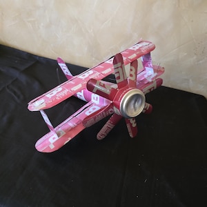 May include: A handmade biplane model crafted from recycled aluminum cans. The plane features a pink and white color scheme, with a silver can used for the engine. The wings and body are detailed with printed text and graphics.