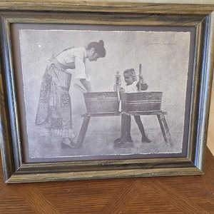 May include: A framed black and white print depicting a woman and child engaged in washing clothes. The woman, wearing a long skirt and apron, is leaning over a washtub. The child is seated in a smaller tub, holding a washboard. The frame is a mix of brown and green.