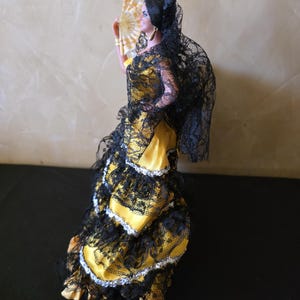 以下が含まれることがあります： フラメンコダンサーのフィギュア。黄色と黒のレースのドレスを着て、扇子を持っています。ダンサーは黒いレースのベールと髪にピンクの花をしています。ドレスは、フリルとレースが特徴です。