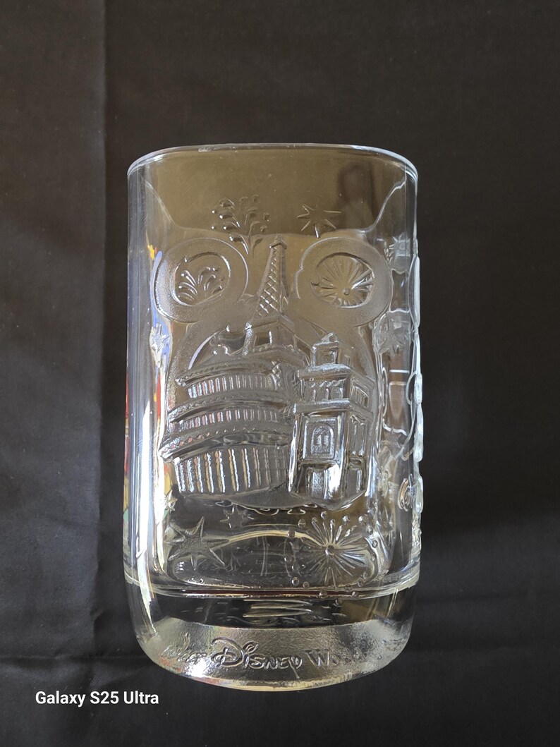 May include: Clear glass tumbler with etched Disney World imagery. The design includes iconic landmarks and fireworks. The base of the glass is inscribed with "Disney World".