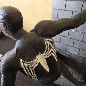K&ouml;nnte beinhalten: Schwarze Figur eines Superhelden in geduckter Haltung, mit einem wei&szlig;en Spinnenemblem auf dem R&uuml;cken. Die Figur ist vor einer grauen Backsteinmauer positioniert, mit einem ausgestreckten Arm. Die Figur ist etwa 15 cm hoch.
