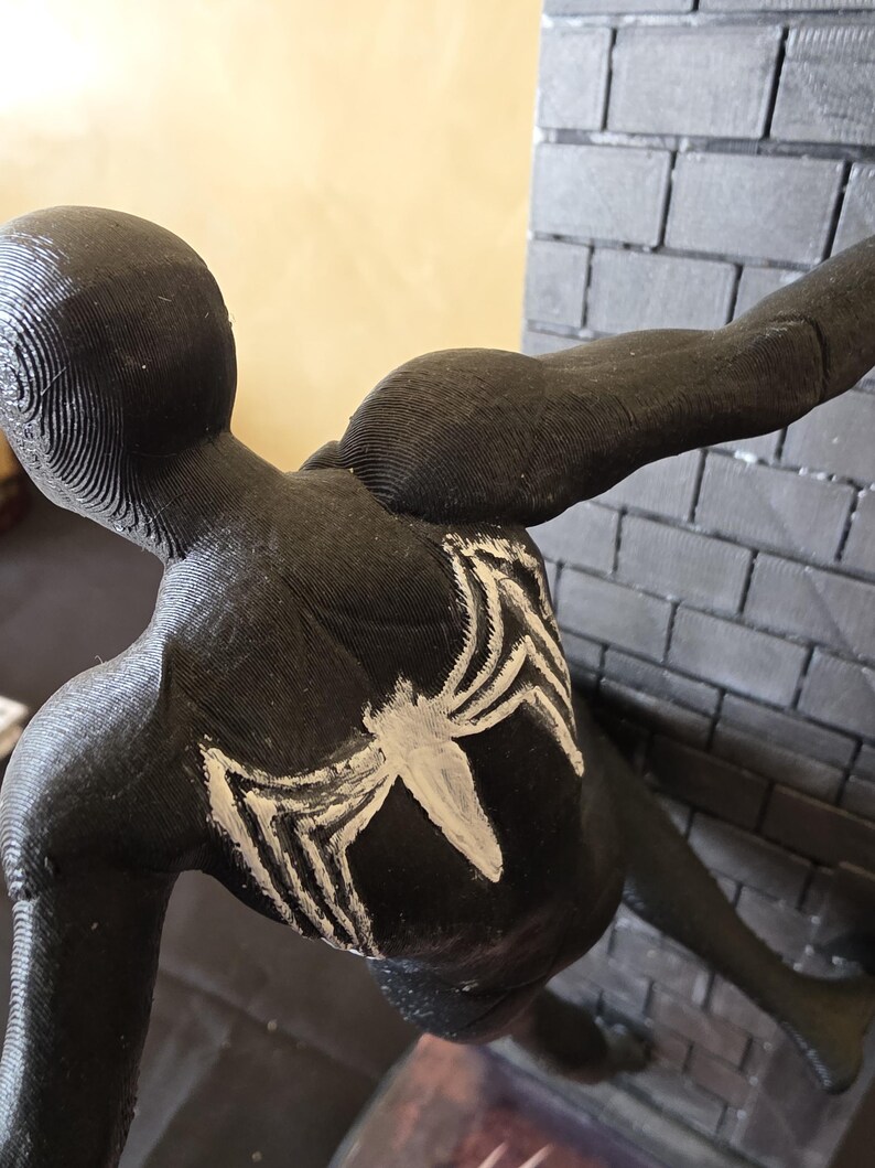 K&ouml;nnte beinhalten: Schwarze Figur eines Superhelden in geduckter Haltung, mit einem wei&szlig;en Spinnenemblem auf dem R&uuml;cken. Die Figur ist vor einer grauen Backsteinmauer positioniert, mit einem ausgestreckten Arm. Die Figur ist etwa 15 cm hoch.