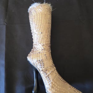 May include: A decorative boot-shaped object covered in gold sequins with a pointed toe and a tall, slender black stiletto heel. The top opening of the boot features a light-colored faux fur trim, and the entire structure is finished with a silver-toned braided cord along the base and seams.
