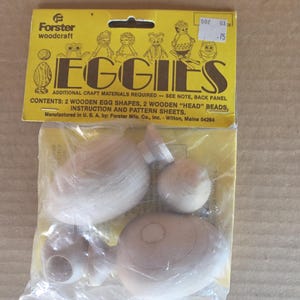 Könnte beinhalten: Eine Packung Holzbastelbedarf mit der Aufschrift "EGGIES" von Forster woodcraft. Die Packung enthält hölzerne Eiformen und Kopfperlen. Die Verpackung ist gelb mit Illustrationen des fertigen Bastelprojekts.